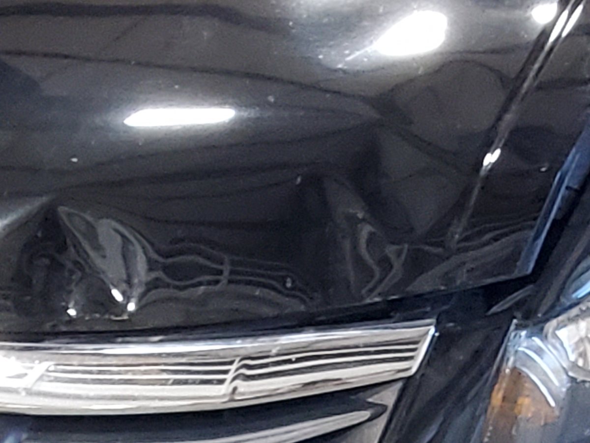 A black Honda Accord in the shop with the front bumper and left fender creased in a collision.