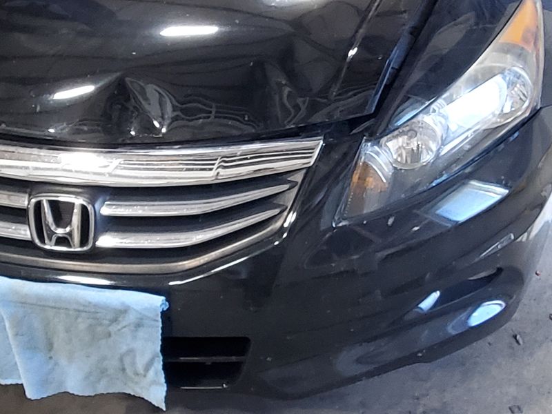 A black Honda Accord in the shop with the front bumper and left fender creased in a collision.